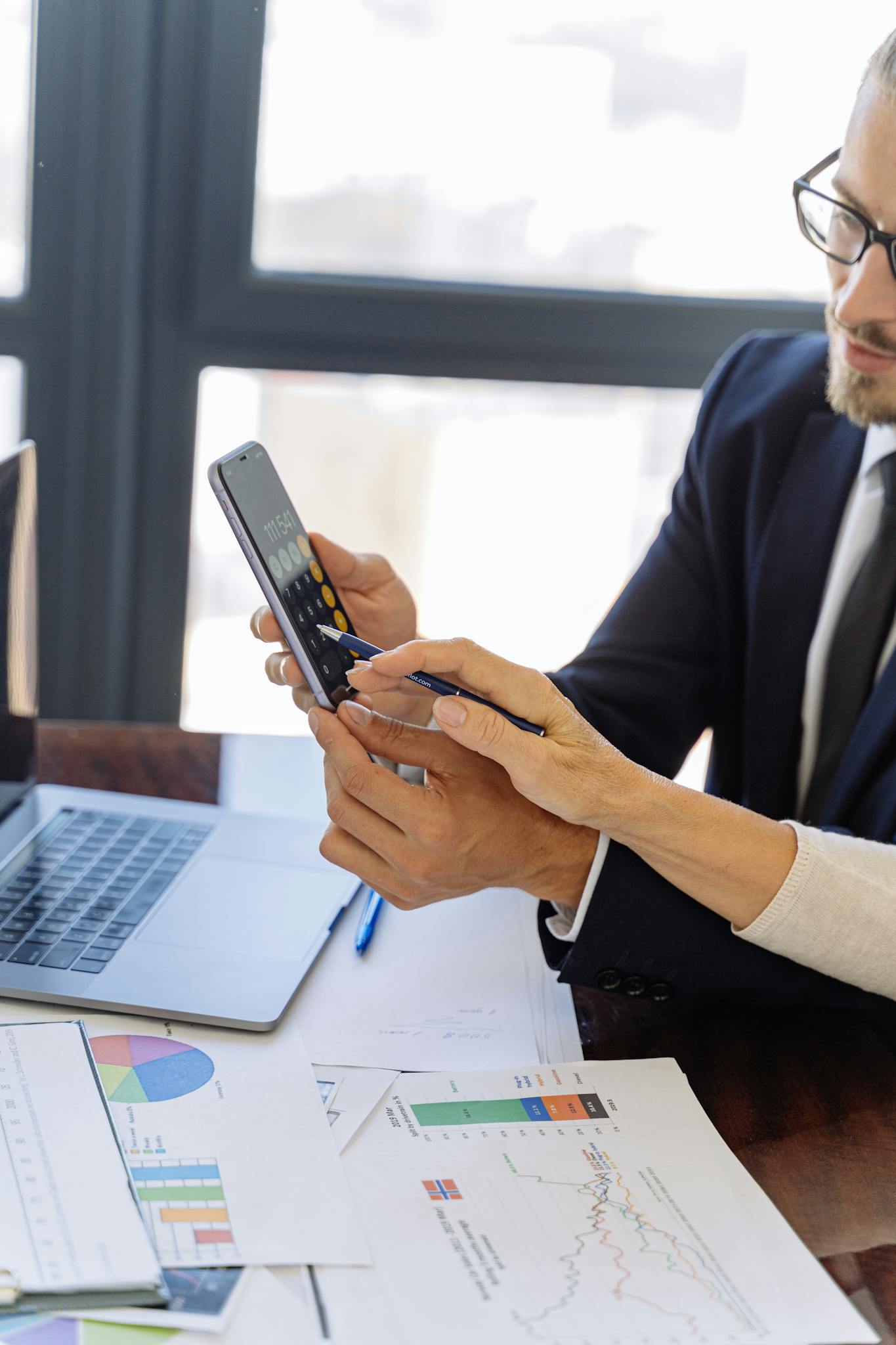 Business professionals reviewing financial reports on a smartphone at a desk with papers and a laptop in an office setting.